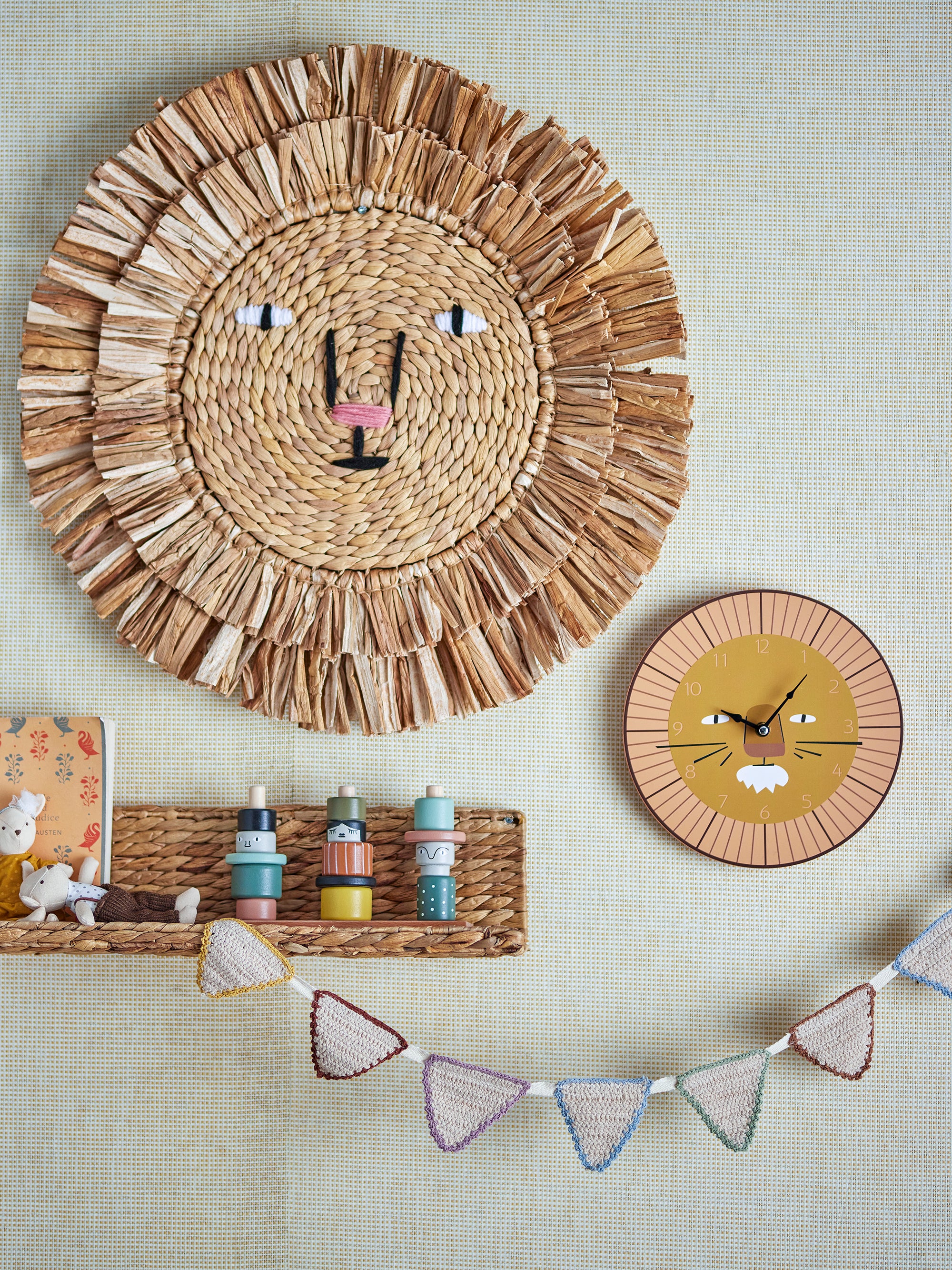 A wall in a children's room with rattan wall decor and a wall clock both resembling an animated lions face. Underneath there is a small rattan shelf decorated with toys and a knitted garland in white.