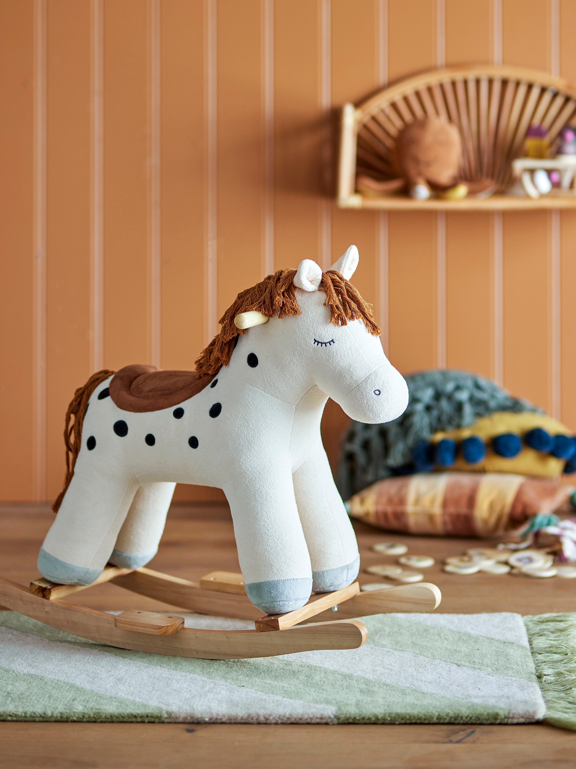 Plush rocking toy shaped as a horse in white with black dots and a brown mane and tail. Placed in a children's room with orange/brown striped walls on a green and white striped rug.