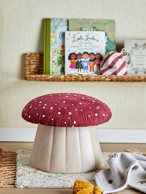 A soft pouf, shaped and colored as a red/white mushroom with dots. Placed in a children's room, with a book rack with children's books behind it.