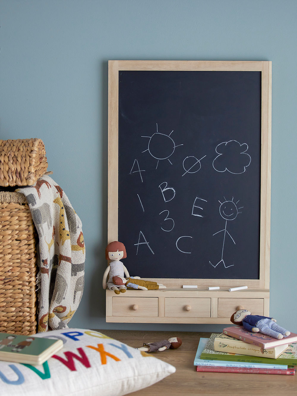 A children's room with a blackboard on a blue wall. The blackboard is decorated with white chalk paintings and is framed in a light tree sort with drawers underneath.